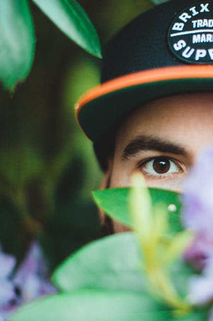 A gardener wearing a wide-brimmed hat and dark clothing is trimming a dense, flowering hedge with long-handled pruning shears. The hedge features lush, green leaves with clusters of pale pink flowers,