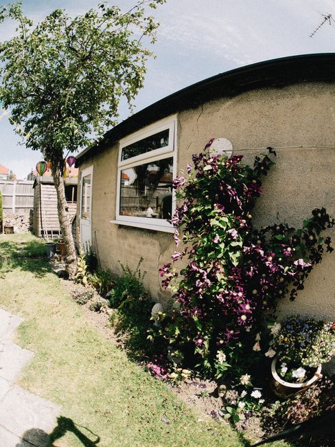A small, curved garden area adjacent to a modest, single-story house with cream-colored exterior walls and large rectangular windows. The garden features a narrow, grassy lawn bordered by a border of