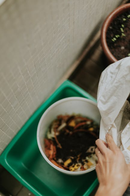 A person's hand is placing organic garden waste, such as vegetable peels and plant debris, into a white compost bin positioned on a green and grey plastic tray, against a beige tiled wall. In the back
